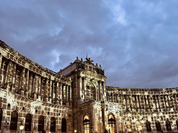 Historic curved palace facade lit at dusk with decorative light projections on columns and arches.