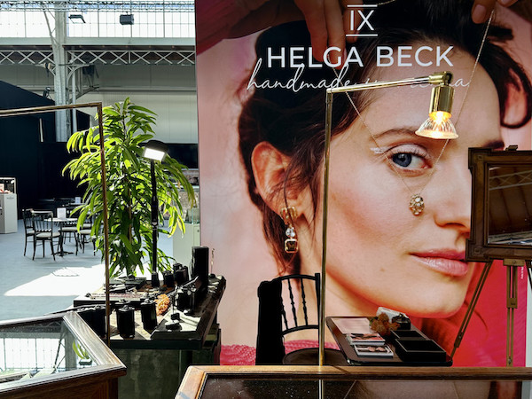 Makeup booth at a trade show; large Helga Beck poster of a woman's face, plus jewelry and makeup displays, a lamp, and a potted plant in the background.
